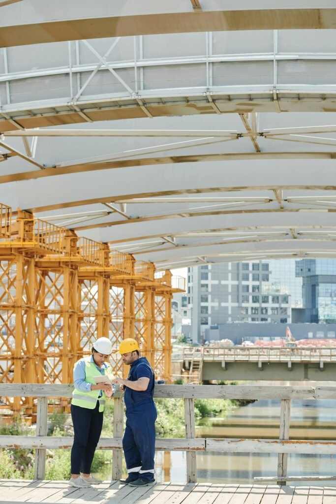 Home Two engineers discussing plans at an outdoor construction site under a bridge.